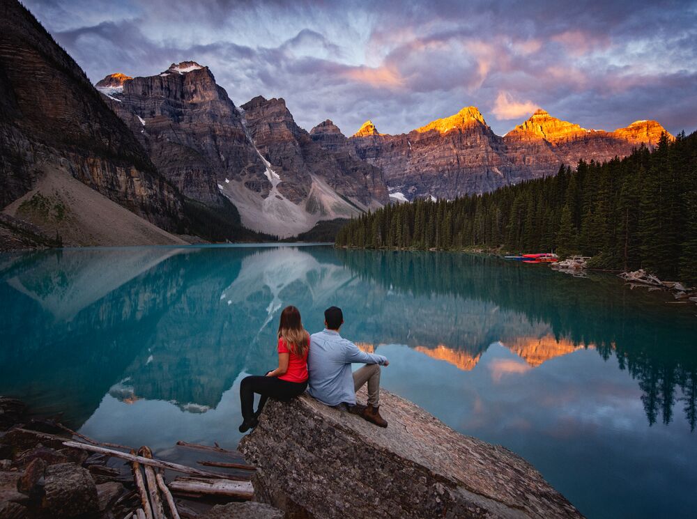 Two people sit on a rock at Moraine Lake at sunrise.